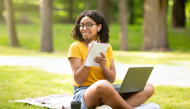 Online education. Happy black girl taking notes while watching web lesson on laptop at park