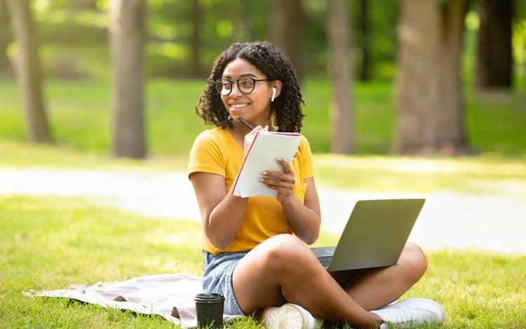 Online education. Happy black girl taking notes while watching web lesson on laptop at park