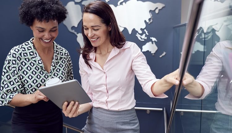 Female Colleagues With Digital Tablet Meeting On Stairs Of Office With World Map In Background