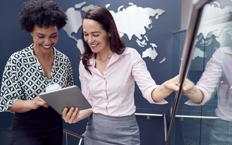 Female Colleagues With Digital Tablet Meeting On Stairs Of Office With World Map In Background