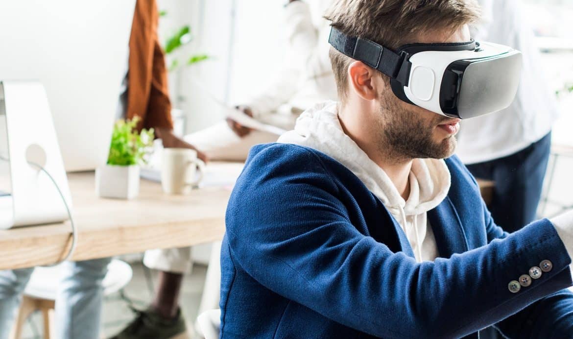 panoramic shot of young businessman touching something with finger while using vr headset in office