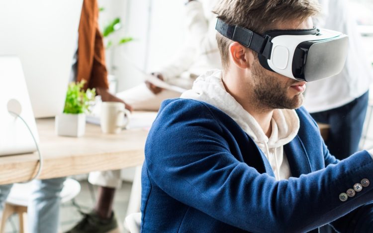 panoramic shot of young businessman touching something with finger while using vr headset in office